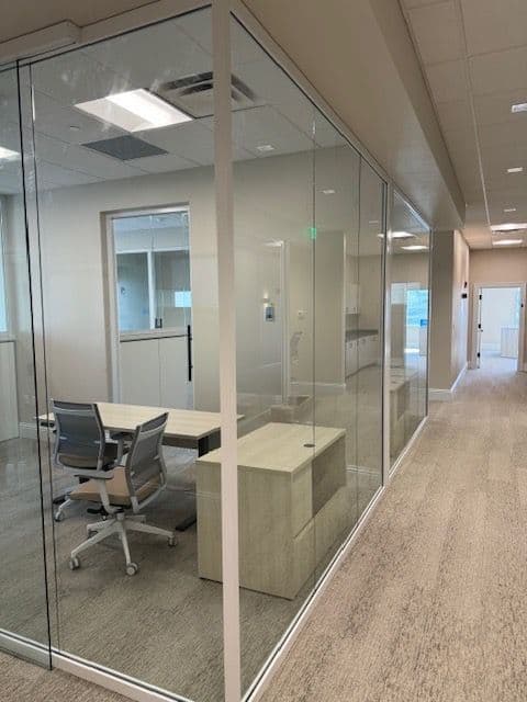 Glass-fronted private office seen from the corridor, with a light-wood desk and return and two grey mesh task chairs